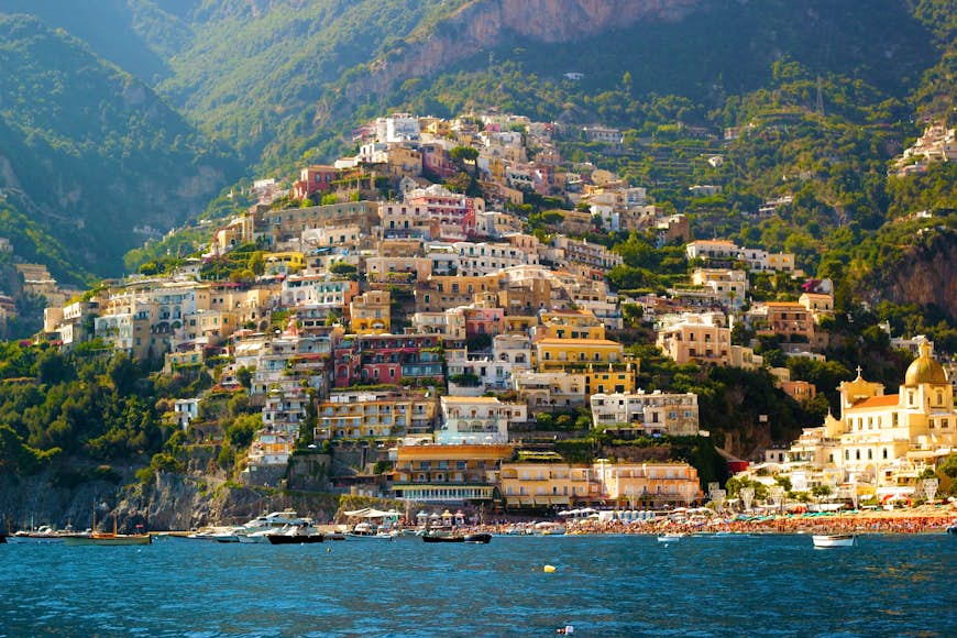 Que ver en Italia Casas coloridas cubren una empinada ladera costera, formando parte de la ciudad de Positano en la costa de Amalfi, Italia. La foto está tomada desde el mar y los barcos están atracados en el agua cerca de la orilla.