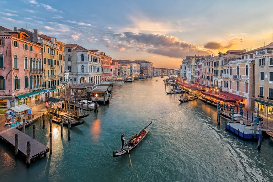 Que ver en Italia. Una góndola flota por un amplio canal en Venecia, Italia, al atardecer. El canal está bordeado de cafés y bares donde la gente se sienta afuera, con botes amarrados al borde del canal.