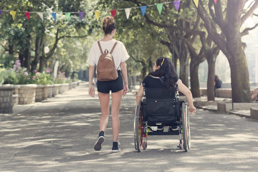 Transporte accesible en Italia ♿Una joven camina junto a su amiga en silla de ruedas en la localidad italiana de Tempio Pausania. El camino es ancho y está flanqueado por vegetación.