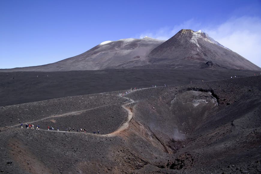 Que ver en Italia.Grupos de excursionistas recorren el camino sinuoso hasta la cumbre ennegrecida del monte Etna en Sicilia, Italia. El volcán negro ceniza se contrasta con un cielo azul claro.