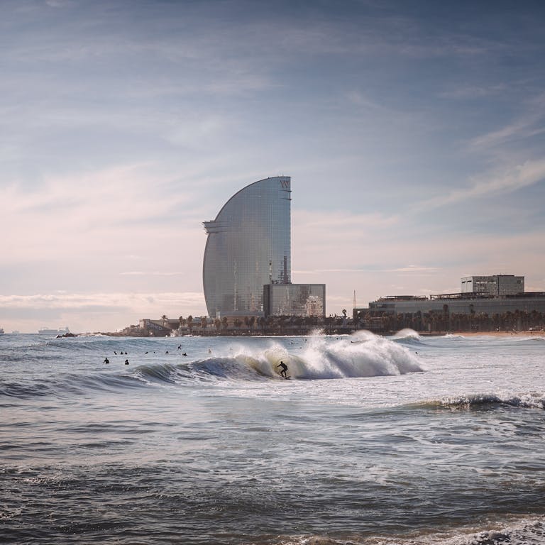 Clima y temperatura de Barcelona, mejor epoca para visitar Barcelona,Surfer navigating waves with the backdrop of the iconic W Barcelona Hotel on a sunny day.